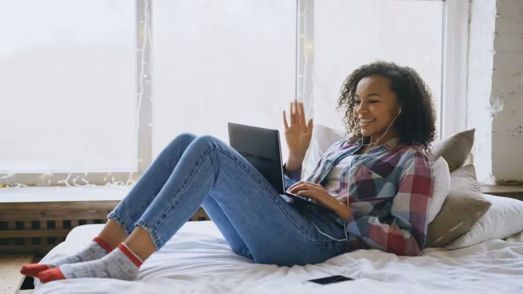 Laptop előtt mosolyogva integető nő az ágyon, aki egy online angol magántanár kezdőknek indított, feszültségmentes órájához csatlakozik.